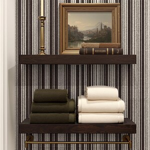 May include: Two dark wooden shelves with stacked towels and decorative items. The top shelf holds a framed landscape painting, books, and a gold candlestick. The bottom shelf displays folded green and cream-colored towels, with a gold towel bar below.