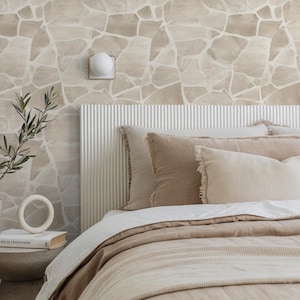 Puede incluir: Una escena de dormitorio con una cama con ropa de cama beige y almohadas decorativas. El cabecero blanco tiene líneas verticales. La pared detrás de la cama tiene un patrón de piedra. Una pequeña mesita de noche sostiene un libro y un objeto blanco en forma de anillo.