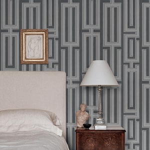 Può includere: Una camera da letto con testiera imbottita beige chiaro, comodino in legno scuro e lampada da tavolo con paralume bianco. Un piccolo busto è appoggiato sul comodino. La parete presenta carta da parati grigia con motivo geometrico.