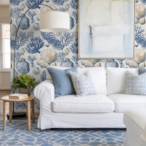 Puede incluir: Un salón con un sofá blanco, cojines azules y una alfombra estampada. La pared presenta un papel pintado con diseño de conchas marinas y corales en azul y beige. Una pintura con tonos azules cuelga sobre el sofá.