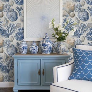 Puede incluir: Una habitaci&oacute;n de tem&aacute;tica costera con un mueble azul claro que contiene jarrones y botes de cer&aacute;mica azules y blancos. Se ve un sill&oacute;n blanco con un coj&iacute;n azul con estampado de olas. El papel pintado presenta conchas marinas y corales.