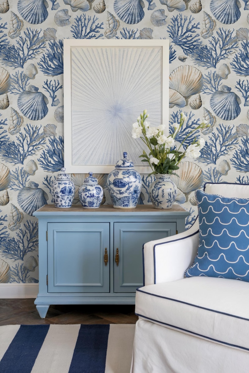 Puede incluir: Una habitaci&oacute;n de tem&aacute;tica costera con un mueble azul claro que contiene jarrones y botes de cer&aacute;mica azules y blancos. Se ve un sill&oacute;n blanco con un coj&iacute;n azul con estampado de olas. El papel pintado presenta conchas marinas y corales.