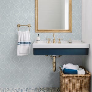 May include: A bathroom scene featuring a gold-framed mirror, a white sink with gold fixtures, and a navy blue base. A white towel with blue stripes hangs on a gold towel rack. The walls have a light blue seashell pattern, and a wicker basket holds towels.