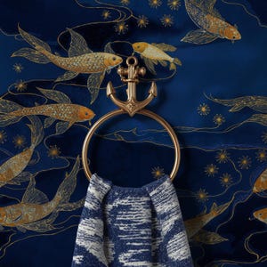 Könnte beinhalten: Ein goldfarbener Handtuchring aus Metall in Ankerform, montiert vor einem dunkelblauen Stoffhintergrund mit goldenen Koi-Karpfen- und Sternmustern. Ein gefaltetes, blau-weißes, strukturiertes Handtuch hängt durch den runden Ring des Ankers.