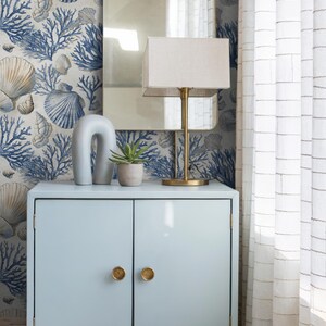 Puede incluir: Un mueble azul claro con pomos dorados, una l&aacute;mpara dorada y un espejo. El papel pintado presenta un dise&ntilde;o de conchas marinas y coral en azul y beige. Una cortina blanca est&aacute; a la derecha.
