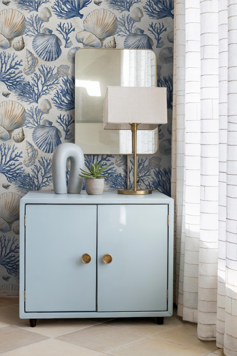 Puede incluir: Un mueble azul claro con pomos dorados, una l&aacute;mpara dorada y un espejo. El papel pintado presenta un dise&ntilde;o de conchas marinas y coral en azul y beige. Una cortina blanca est&aacute; a la derecha.