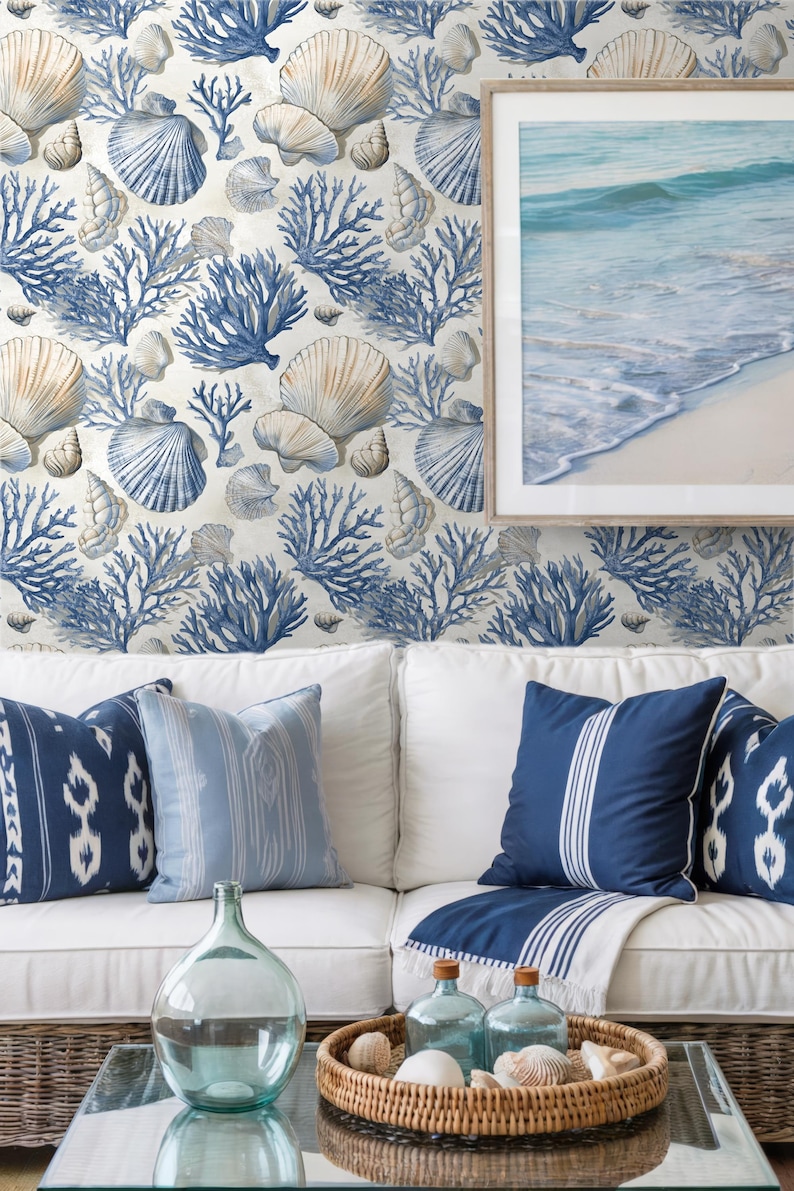 Puede incluir: Sal&oacute;n de tem&aacute;tica costera con un sof&aacute; blanco con cojines azules estampados y una mesa de centro de mimbre. La pared est&aacute; decorada con papel pintado de conchas marinas y corales. Una imagen enmarcada del mar y la playa est&aacute; en la pared.