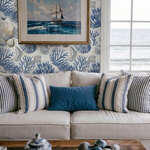 Puede incluir: Sal&oacute;n de estilo costero con un sof&aacute; blanco con cojines a rayas y lisos azules. Un cuadro enmarcado de un barco cuelga en una pared con papel pintado de conchas marinas y corales. Una mesa de centro de madera exhibe conchas decorativas.