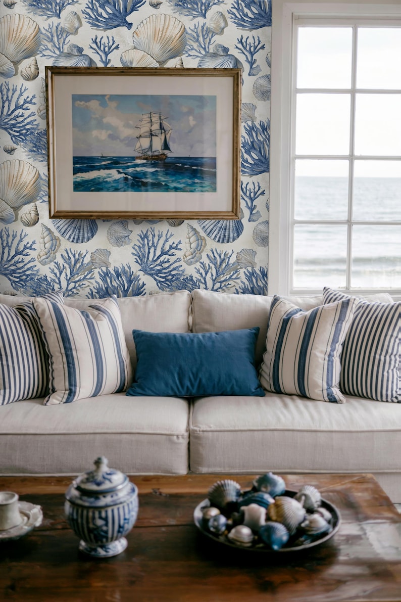 Puede incluir: Sal&oacute;n de estilo costero con un sof&aacute; blanco con cojines a rayas y lisos azules. Un cuadro enmarcado de un barco cuelga en una pared con papel pintado de conchas marinas y corales. Una mesa de centro de madera exhibe conchas decorativas.
