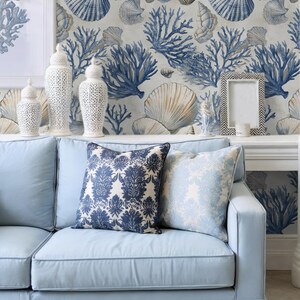 Puede incluir: Sof&aacute; azul claro con dos cojines decorativos con estampados florales. El fondo muestra un papel pintado con motivos de conchas marinas y coral. Jarrones de cer&aacute;mica blanca y un marco en una estanter&iacute;a.