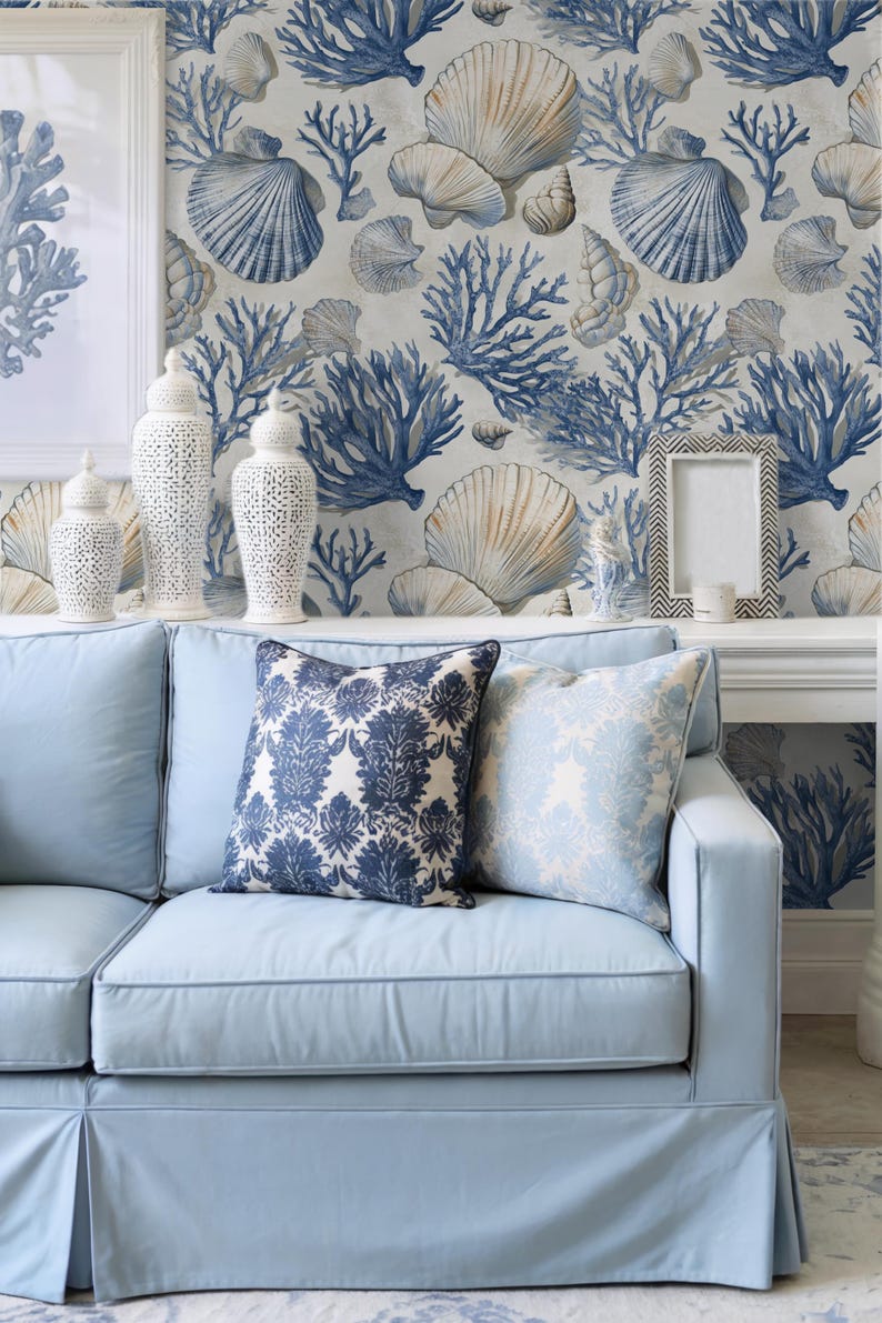 Puede incluir: Sof&aacute; azul claro con dos cojines decorativos con estampados florales. El fondo muestra un papel pintado con motivos de conchas marinas y coral. Jarrones de cer&aacute;mica blanca y un marco en una estanter&iacute;a.