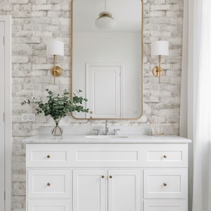 May include: A white bathroom vanity with gold fixtures and a marble worktop. A large gold-framed mirror hangs above the vanity, reflecting a white door and a pendant light. The wall is covered in white brick wallpaper.