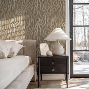 May include: A bedroom scene featuring a bed with white linens, a dark nightstand with two drawers, and a white lamp. The wall has a beige and brown wavy pattern. A window with a view of trees is visible.