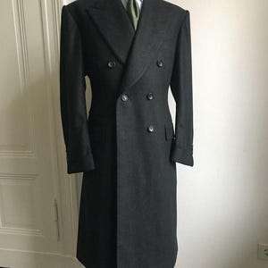 Puede incluir: Un abrigo cruzado gris antracita con seis botones, exhibido en un maniqu&iacute;. El abrigo presenta solapas con muescas, dos bolsillos delanteros y una silueta larga y entallada. Se ven una camisa blanca y una corbata verde oliva.