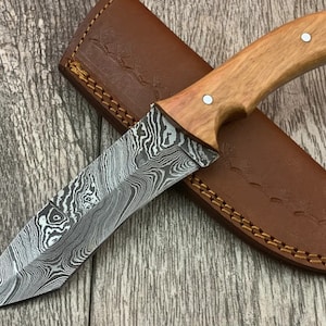 May include: A Damascus steel hunting knife with a wooden handle and a leather sheath. The blade has a wavy pattern and the handle is a light brown colour.
