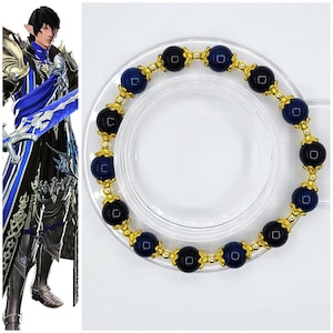 Könnte beinhalten: Ein goldenes Armband mit abwechselnd schwarzen und blauen Perlen. Das Armband liegt auf einer weißen Oberfläche.
