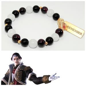 Puede incluir: Una pulsera de cuentas con cuentas blancas, negras y granates alternas, con espaciadores dorados. Se adjunta un dije rectangular dorado con la palabra "REMEMBER". La pulsera se muestra sobre un fondo blanco.