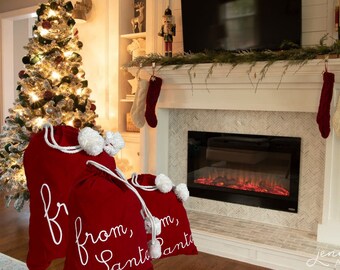 May include: Red velvet Santa sacks with white drawstring closures and fluffy white pom-poms. The sacks have the words "from, Santa" written in white script. A Christmas tree and fireplace are in the background.
