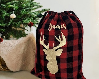 May include: Red and black buffalo plaid Christmas gift bag with a gold deer head silhouette and the name "James" in gold script. The drawstring bag is a festive holiday decoration.