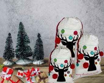 May include: Three white Christmas gift bags with a black deer head design, red pom-poms, and colorful ornaments. The bags have a drawstring closure and the text "merry christmas my deer". Miniature Christmas trees and wrapped gifts are in the background.