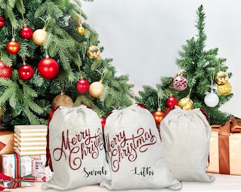 May include: Three off-white Christmas gift bags with red glitter "Merry Christmas" script and personalized names. Each bag has a red ribbon drawstring. The bags are in front of a decorated Christmas tree with red and gold ornaments.
