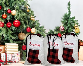 May include: Three white Christmas gift bags with black drawstrings, each featuring a red and black buffalo plaid stocking design. The bags are personalized with the names "Emma", "Chloe", and "Anna" in red script.