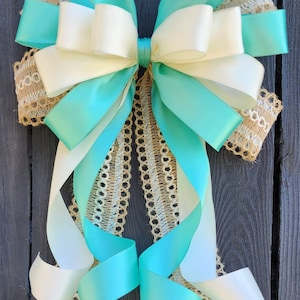May include: A large turquoise and ivory bow with burlap and lace trim. The bow is tied with a single loop and has long tails. The bow is tied to a wooden surface.