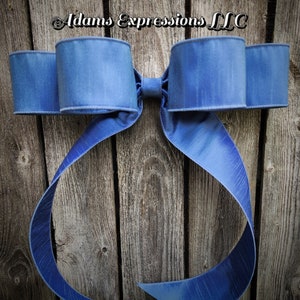 May include: A large, blue decorative bow with a textured appearance. The bow is made of a wide ribbon and features multiple loops and long, flowing tails. The text "Adams Expressions LLC" is visible at the top.