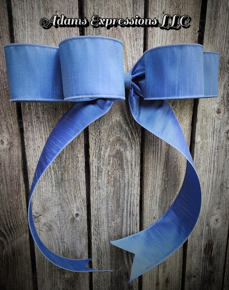 May include: A large, light blue decorative bow with a textured appearance. The bow features three loops on each side and long, flowing tails. The background is a rustic wooden surface. The text "Adams Expressions LLC" is at the top.