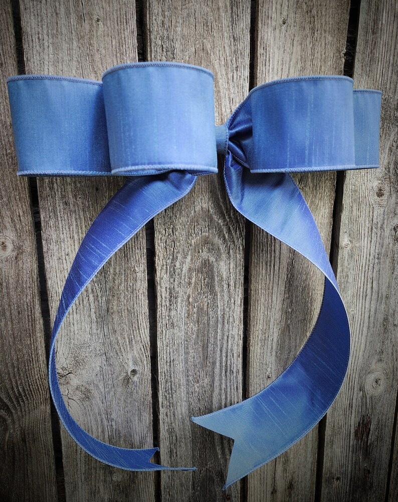 May include: A large, light blue decorative bow with a textured appearance. The bow is made of a wide ribbon and has two large loops on each side, with long, flowing tails. The bow is set against a rustic wooden background.