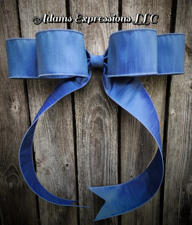 May include: A large, blue decorative bow with a textured finish. The bow is made of a wide ribbon and has multiple loops and long, flowing tails. The bow is mounted on a weathered wooden background. The text "Adams Expressions LLC" is in the upper left corner.
