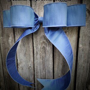 May include: A large, blue decorative bow with long, flowing ribbons. The bow is made of a textured fabric and is mounted on a weathered wooden background. The text "Adams Expressions LLC" is at the top.