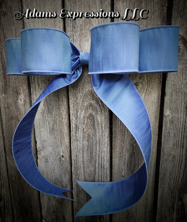 May include: A large, blue decorative bow with long, flowing ribbons. The bow is made of a textured fabric and is mounted on a weathered wooden background. The text "Adams Expressions LLC" is at the top.