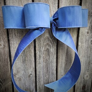 May include: A large, decorative bow in shades of blue, with a textured appearance. The bow is made of a wide ribbon and is displayed against a weathered wooden background. The bow's loops and tails create a classic, elegant design.