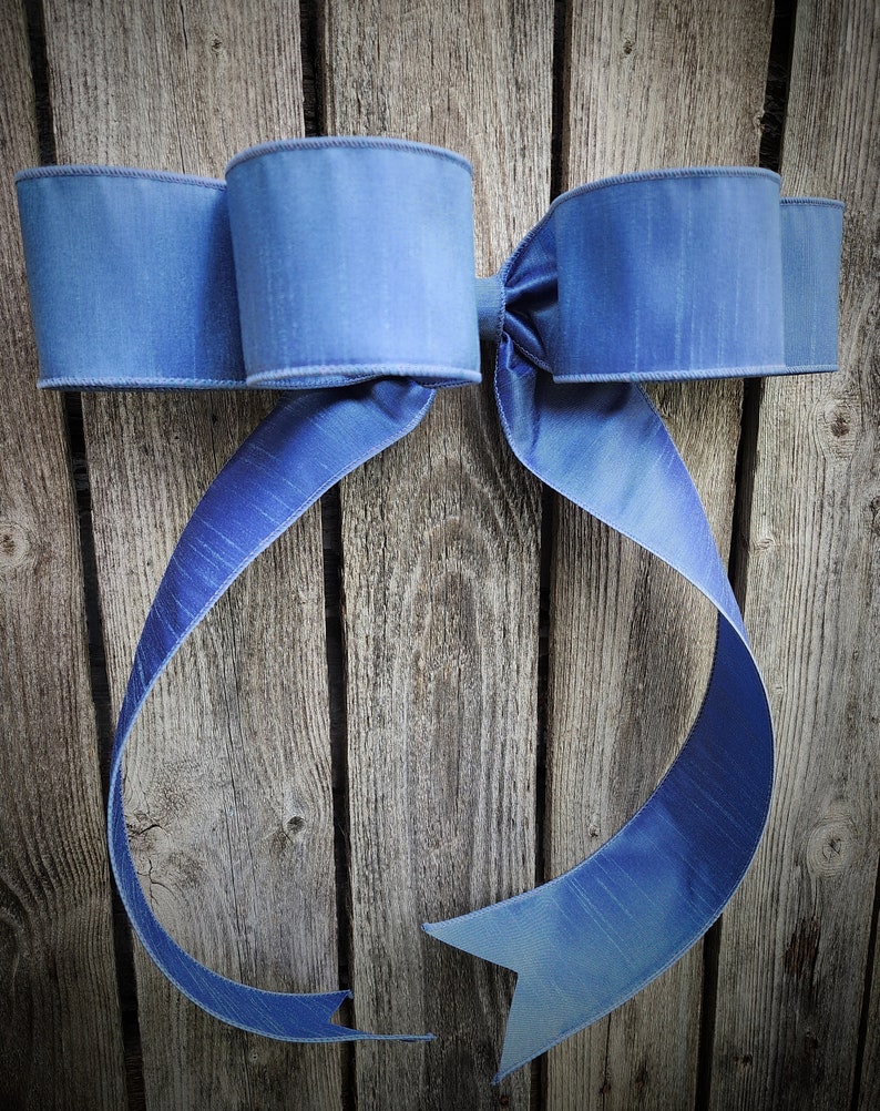 May include: A large, decorative bow in shades of blue, with a textured appearance. The bow is made of a wide ribbon and is displayed against a weathered wooden background. The bow's loops and tails create a classic, elegant design.