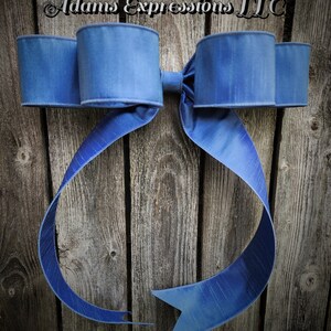 May include: A large, blue decorative bow with a textured finish. The bow features two large loops on each side and long, flowing tails. The background is a rustic wooden surface. The text "Adams Expressions LLC" is visible at the top.