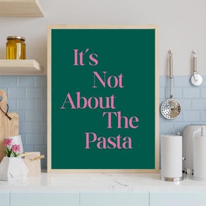 Könnte beinhalten: Ein gerahmter Kunstdruck mit dem Text "It's Not About The Pasta" in rosa auf dunkelgrünem Hintergrund. Der Druck steht auf einer Küchenarbeitsplatte mit Küchenutensilien und Dekoration.