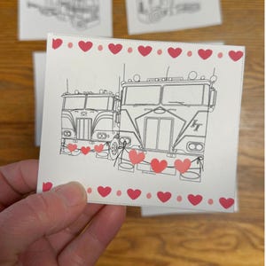 May include: A white card with a black and white illustration of two semi-trucks, decorated with red hearts. The card's border is adorned with small red hearts. The card is held by a hand.