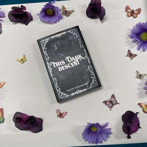 May include: A black book titled "This Dark Descent" by Kalyn Josephson, surrounded by purple flowers and butterfly decorations. The book has a decorative border and is centered on a white surface.