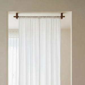 May include: A white curtain hangs from a wooden rod with dark brown brackets, creating a room divider. The curtain is in front of a window, with a chair, side table, and books visible in the background. The room has hardwood floors.