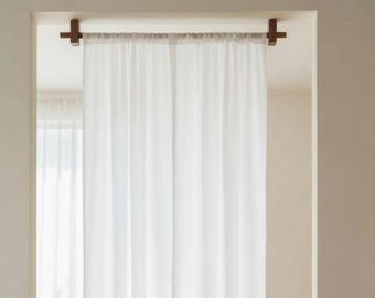 May include: A white curtain suspended from a wooden rail with dark brown brackets, acting as a room divider. The curtain is in front of a window, with a chair, side table, and books in the background. The room has wooden flooring.
