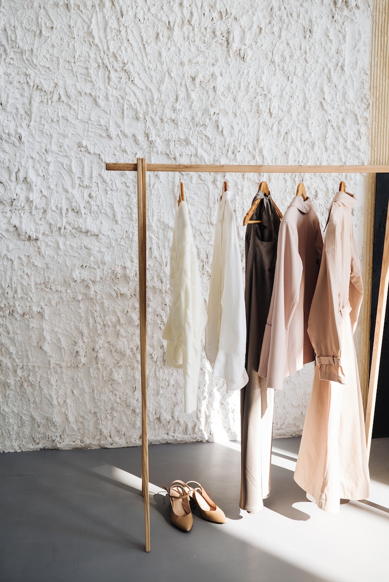 May include: A wooden clothing rack displays several garments: a cream-colored blouse, a white blouse, brown pants, a pink coat, and a light pink trench coat. Beige heels sit on the floor. The background is a textured white wall.