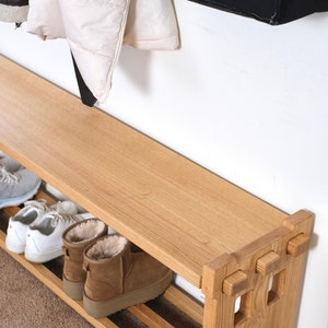 May include: A light-toned wooden shoe bench featuring a flat top surface and a lower slatted shelf for storage. The bench has a sturdy frame with square cut-out details on the side panels. Several pairs of shoes are placed on the lower shelf.
