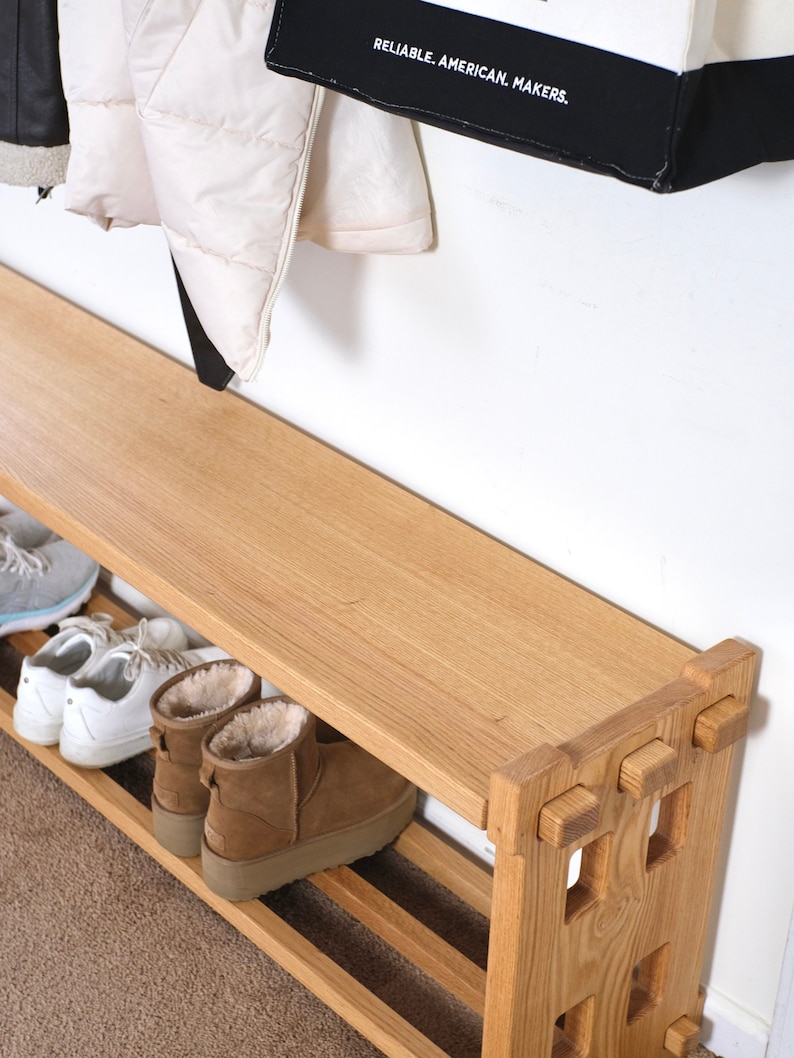 May include: A light-toned wooden shoe bench featuring a flat top surface and a lower slatted shelf for storage. The bench has a sturdy frame with square cut-out details on the side panels. Several pairs of shoes are placed on the lower shelf.