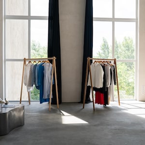 May include: Two wooden clothing racks displaying a variety of shirts, dresses, and blouses. The garments are in shades of white, blue, black, and red. The racks are in a bright room with large windows and black curtains.