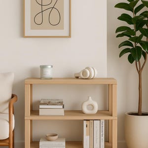 May include: A light wood three-tier shelf unit with decorative items. A framed abstract art print hangs above the shelf. Items on the shelves include a candle, books, a vase, and a decorative sculpture. A potted plant is to the right.