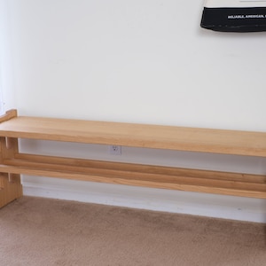 May include: A light-coloured wooden bench featuring a rectangular top surface and a lower shelf for storage. The bench is constructed with sturdy side supports that have a notched, interlocking design, showcasing the natural grain of the wood.