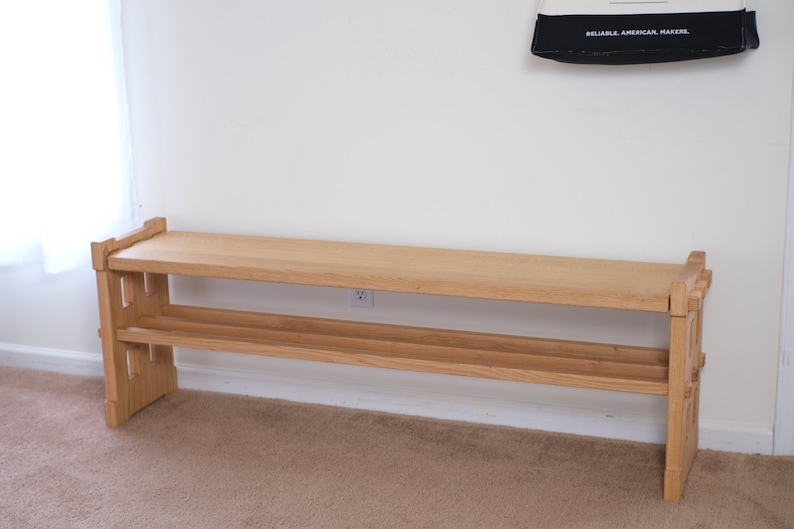 May include: A light-coloured wooden bench featuring a rectangular top surface and a lower shelf for storage. The bench is constructed with sturdy side supports that have a notched, interlocking design, showcasing the natural grain of the wood.