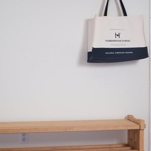 May include: A cream and navy blue canvas tote bag with black handles, featuring the text "HUBBARDTON FORGE" and "RELIABLE. AMERICAN. MAKERS." printed on the front, hanging from a wooden wall-mounted coat rack above a wooden bench.