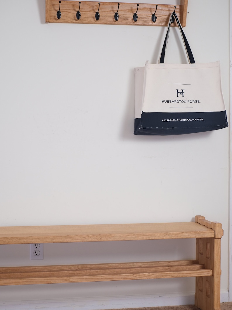 May include: A cream and navy blue canvas tote bag with black handles, featuring the text "HUBBARDTON FORGE" and "RELIABLE. AMERICAN. MAKERS." printed on the front, hanging from a wooden wall-mounted coat rack above a wooden bench.